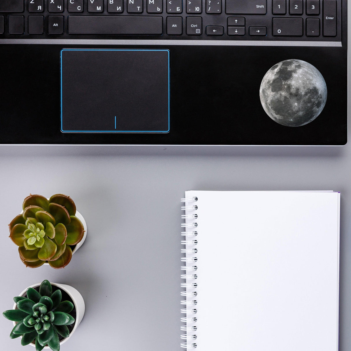 Lunar Luminescence laptop decal featuring a glowing moon design on a black laptop with plants and a notebook.