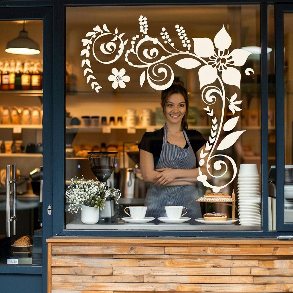 Barista in a café smiling behind a window with decorative vinyl floral decal, showcasing a warm and inviting atmosphere.