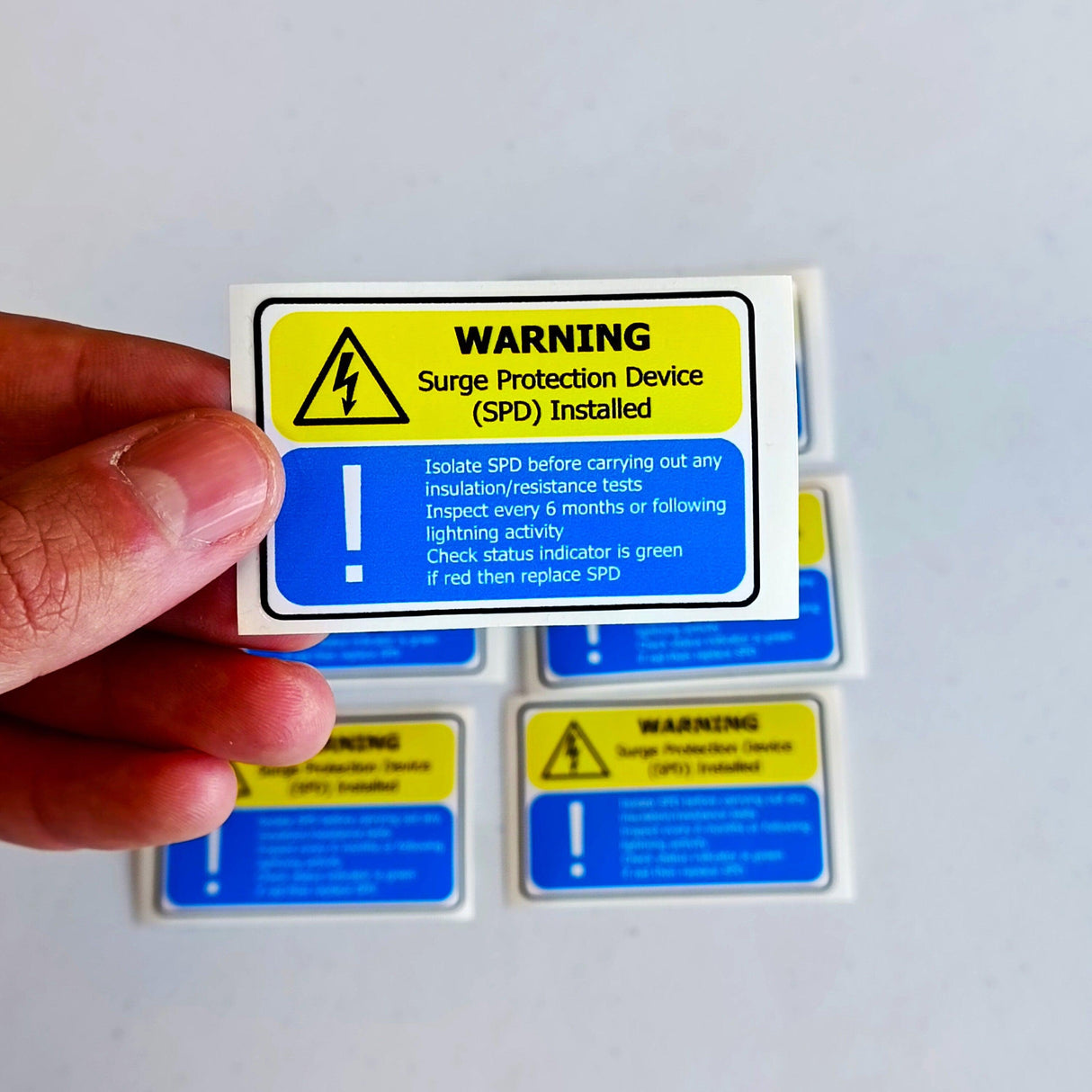 Hand holding yellow and blue Surge Protection Device (SPD) warning label with safety instructions for electrical installations.