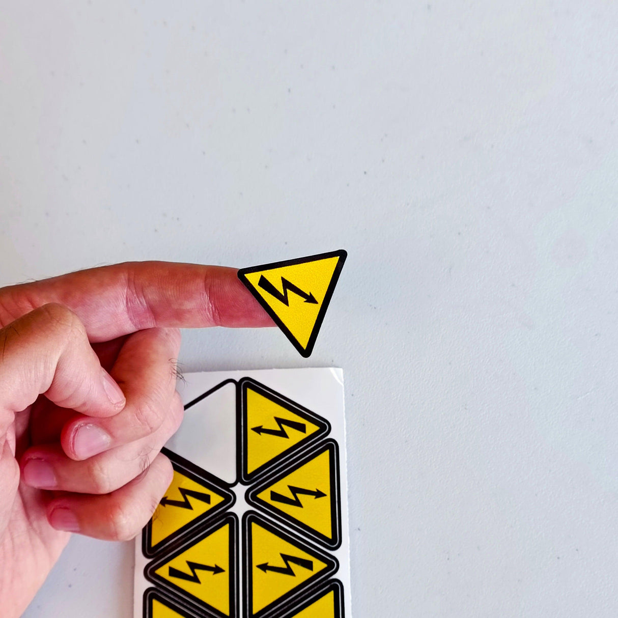 Hand holding a high voltage electrical shock hazard sticker in a yellow triangle shape.