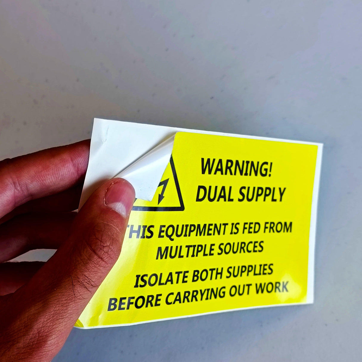 Hand holding a bright yellow dual supply warning label sticker for electrical safety, featuring clear isolation instructions.