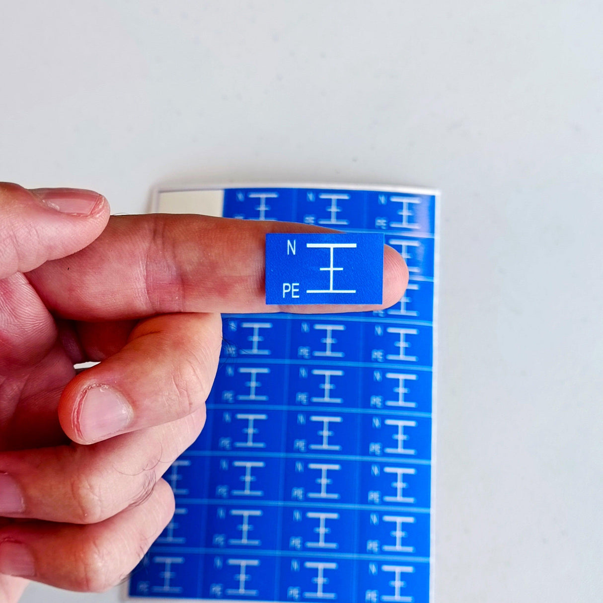 Hand holding blue grounding symbol sticker labeled N and PE above a sheet of similar electrical component labels.