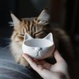 Hand holding a cute 3D-printed cat bowl with a minimalist design, featuring a cat face, in front of a cat.