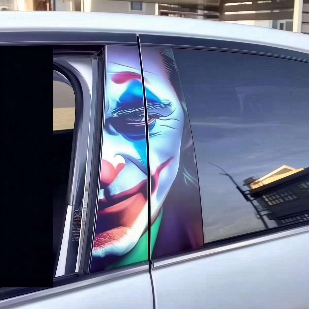 A vibrant Joker face B-pillar car sticker installed on a silver car, showing intense makeup with blue, red, and white tones stretched across the window frame for dramatic visual impact.