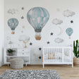 A dreamy nursery room with hot air balloon wall decals in soft blue tones, surrounded by watercolor clouds, stars, and a crescent moon. The wall art complements the white crib and wooden floor in a modern, cozy setting.
