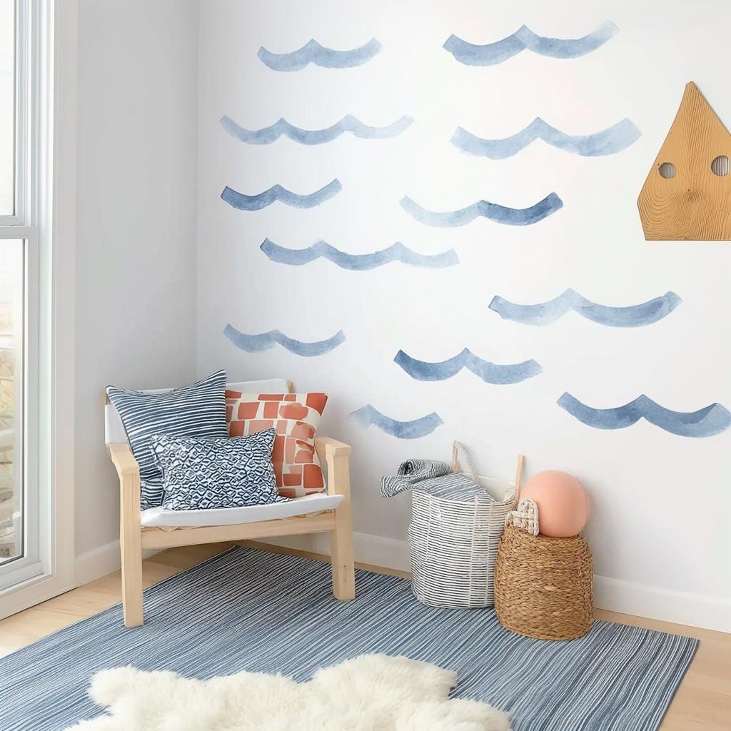 Watercolor wave wall decals in varying shades of blue arranged on a white wall above a cozy chair and storage baskets in a coastal-themed kids' room.