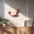3D printed wall shelf in the shape of a cat, displayed in pink above a wooden cabinet with plants and books, modern minimalist room decor