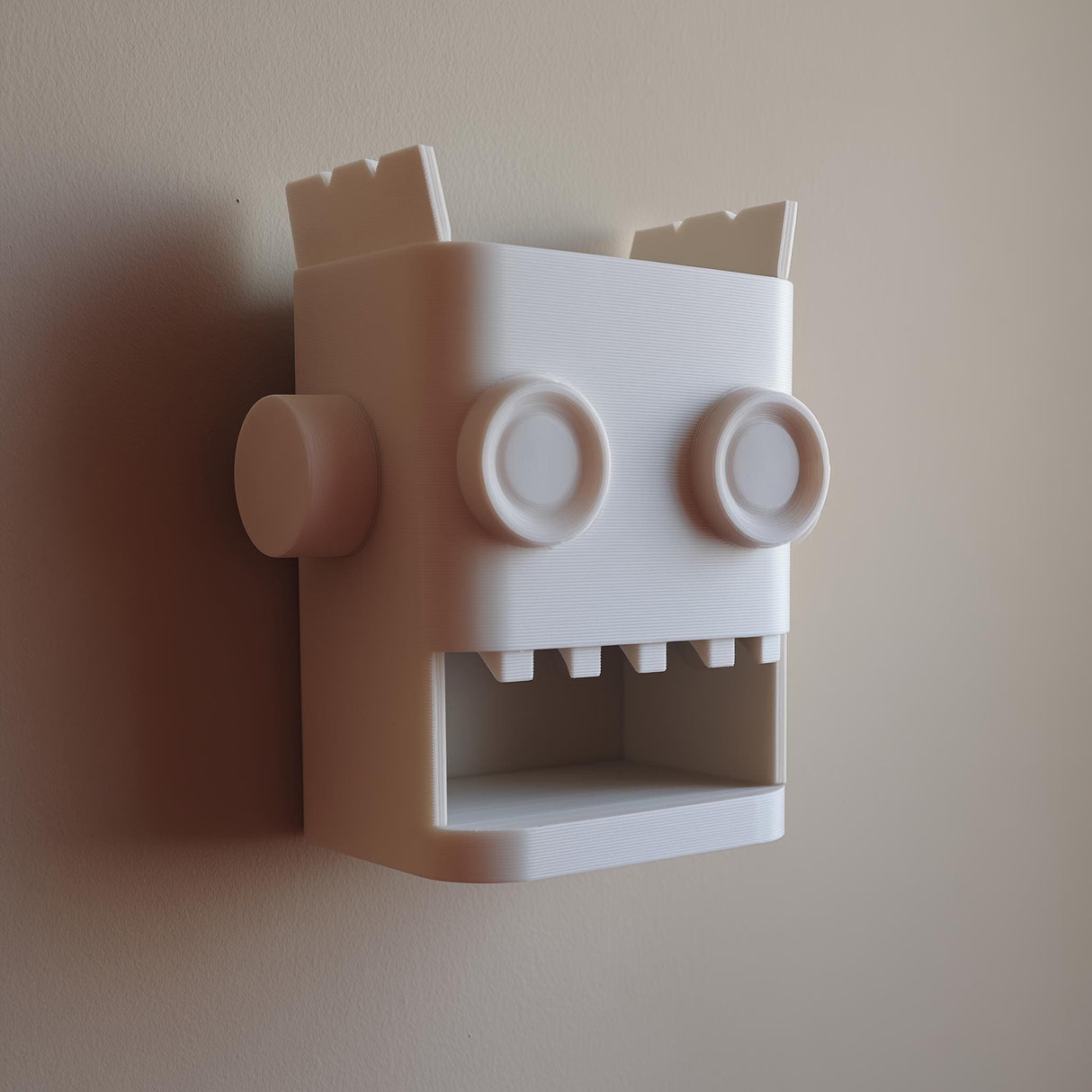 White 3D printed robot head wall organizer with an open mouth storage niche, made of matte PLA and mounted on a plain wall in soft natural light.