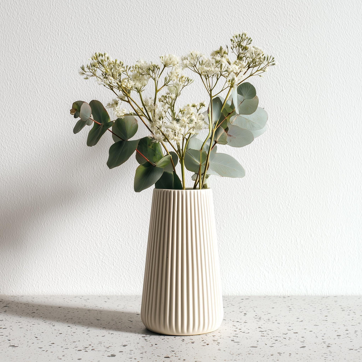 A tall, cream-colored 3D printed origami vase with vertical ribbed lines, holding white dried flowers and eucalyptus stems, placed on a speckled countertop.