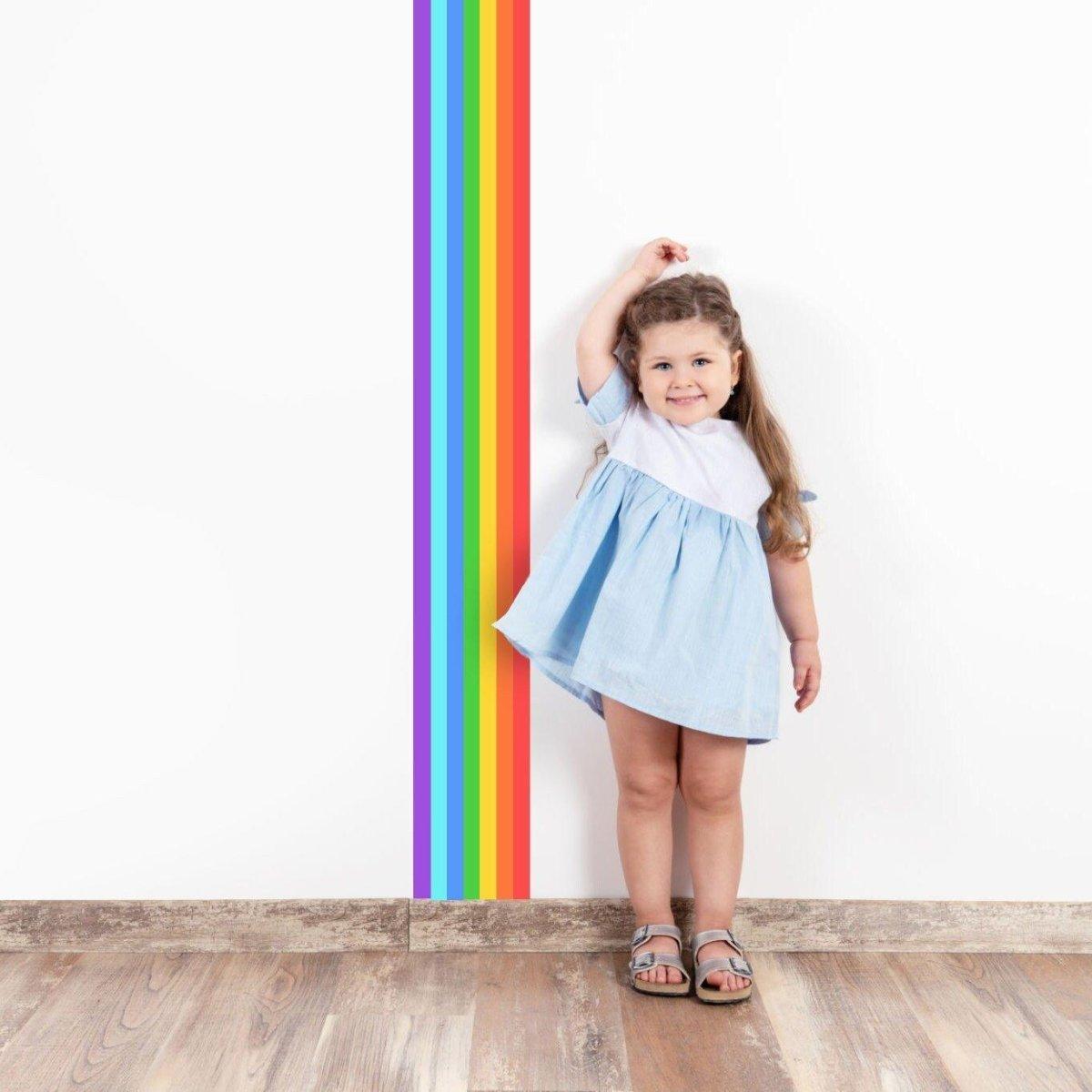 Young girl with a rainbow stripe decal on the wall, wearing a blue dress, smiling happily in a bright, colorful room.