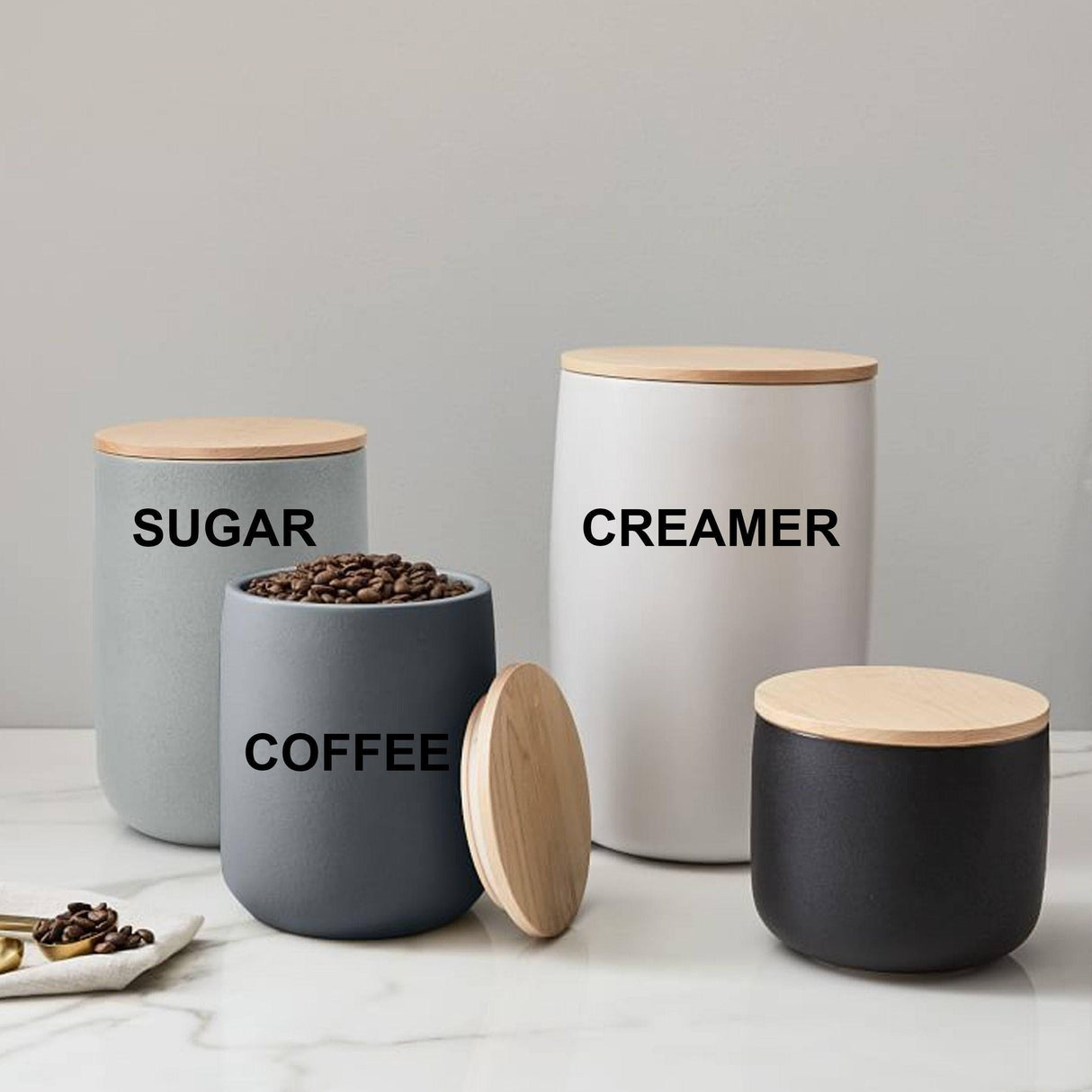 Set of modern storage canisters labeled Sugar, Creamer, Coffee with wooden lids on a marble surface.