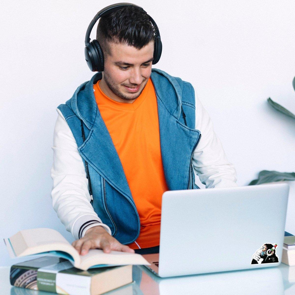 A person wearing headphones using a laptop with a contemplative monkey decal, surrounded by books.
