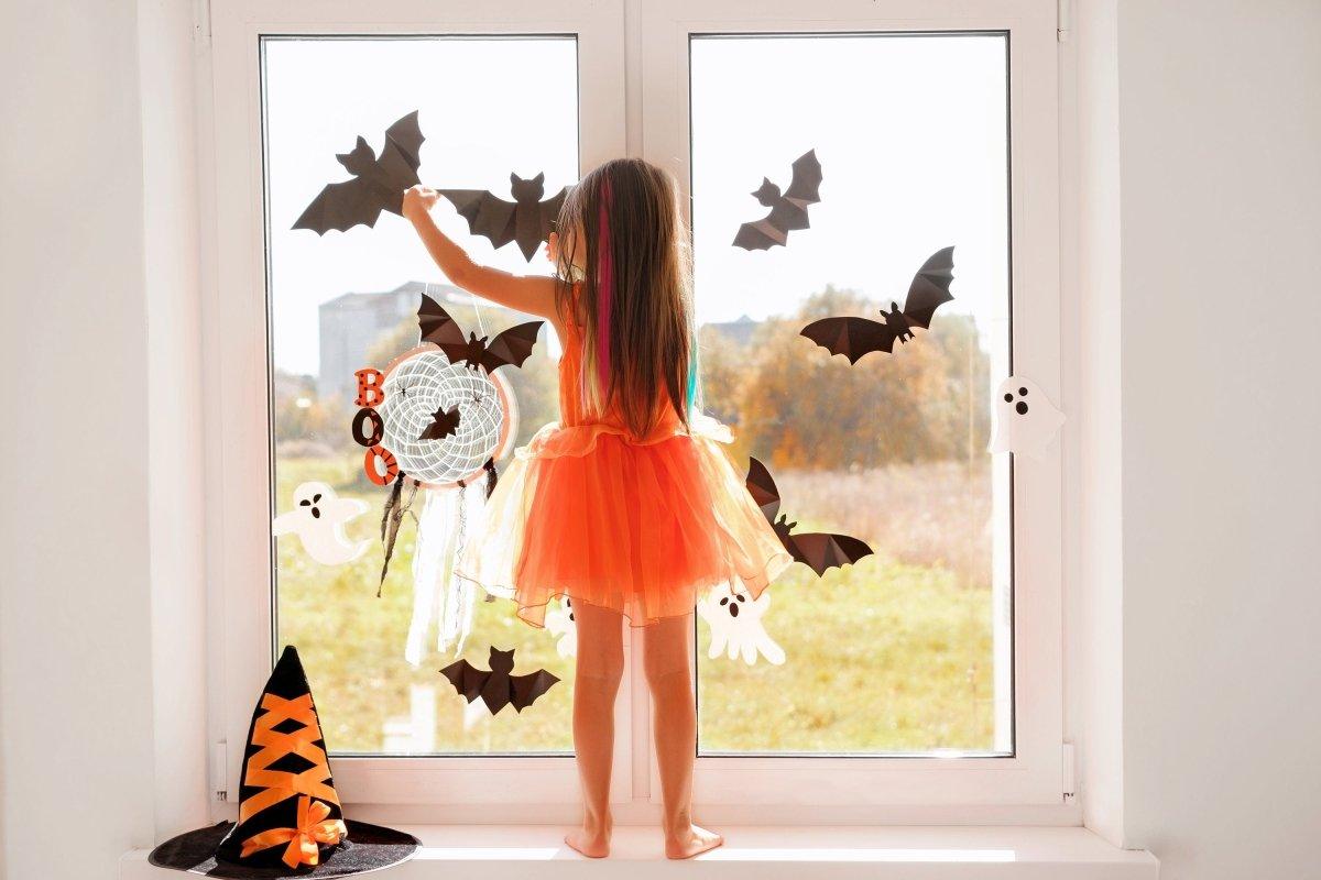 Child decorating a window with Halloween bat decals and spooky decorations in a vibrant orange dress.
