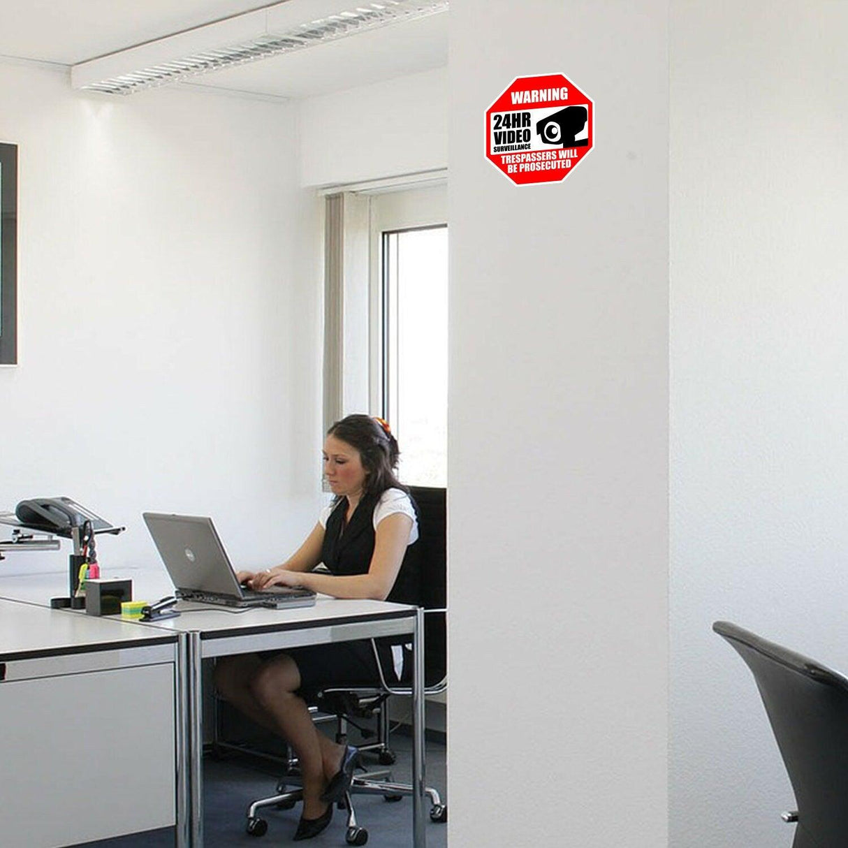 Woman working at desk in office next to a 24-hour video surveillance warning sign decal.