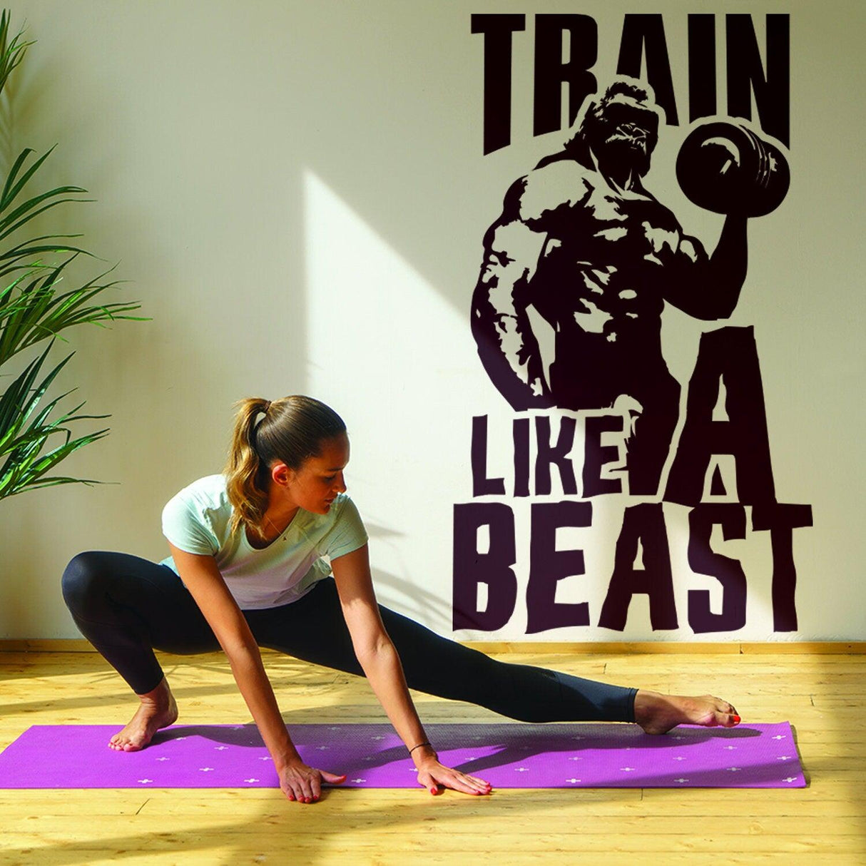 Woman stretching on a yoga mat with a 'Train Like A Beast' wall decal in a bright exercise room.