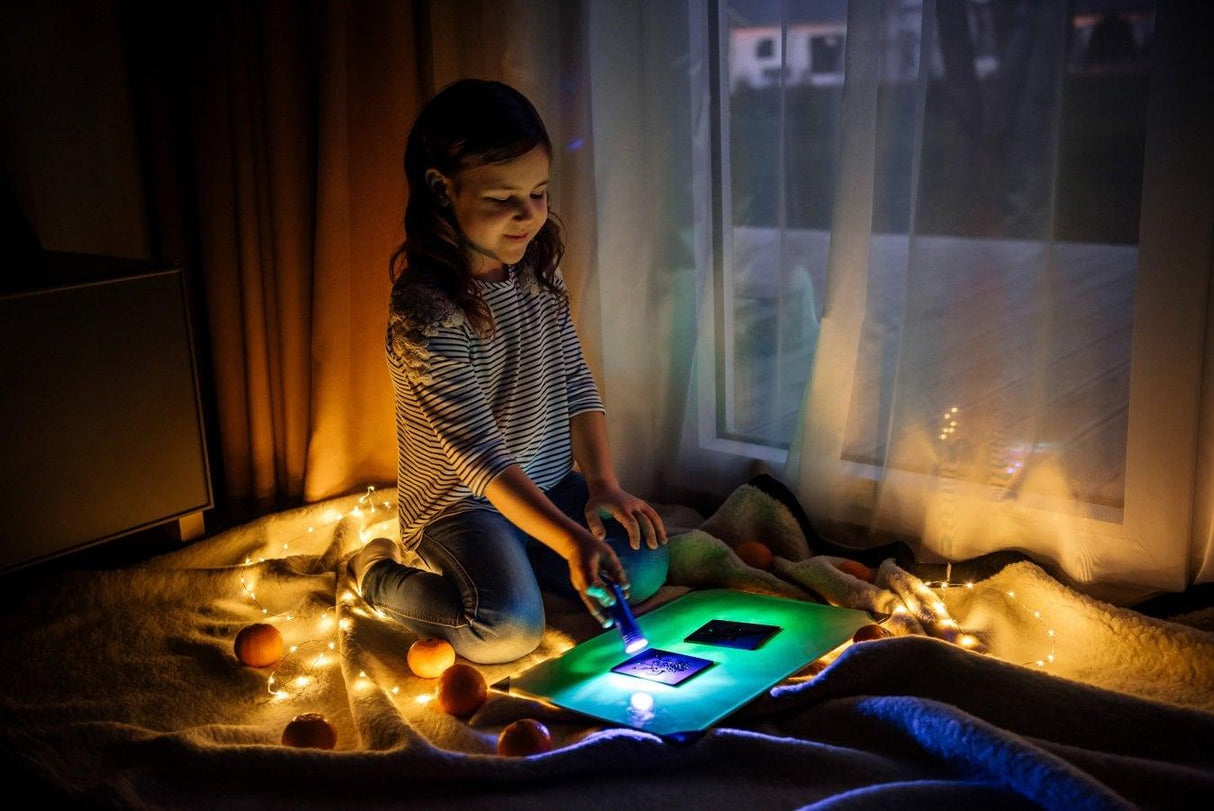 A child using a glow in the dark drawing board, creating neon art in a cozy room with string lights.