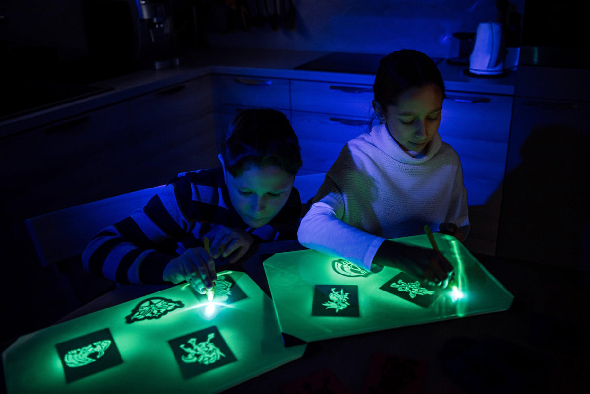 Children drawing on neon effect light drawing boards in a dark room, showcasing their glowing artwork.