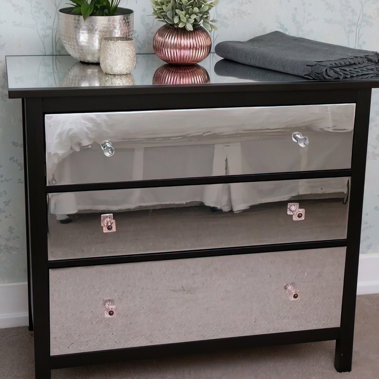 Mirrored black dresser with three drawers, decorative plants, and a cozy blanket on top.