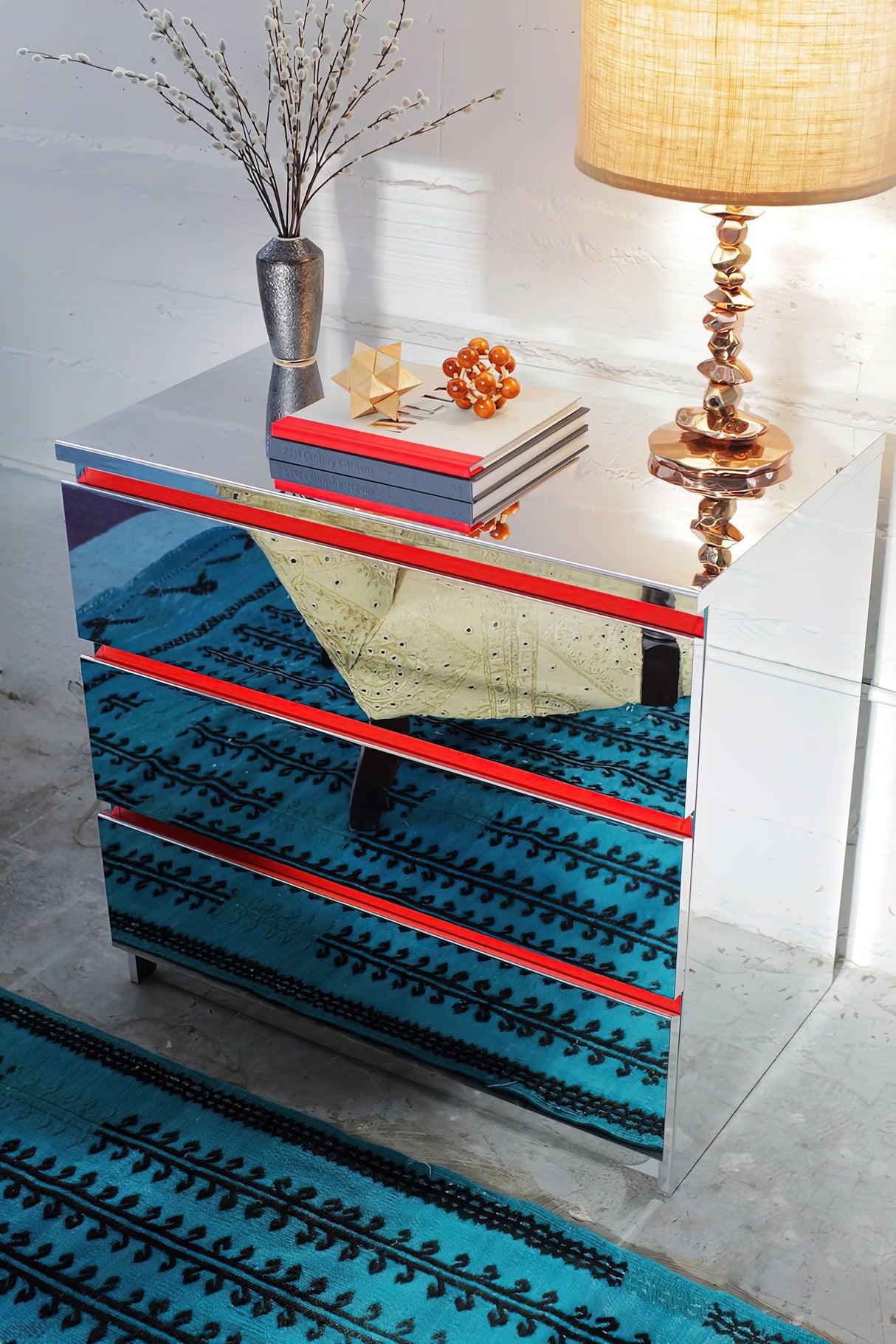 Stylish mirrored dresser with red accents, lamp, vase, and decor on a vibrant blue patterned rug.