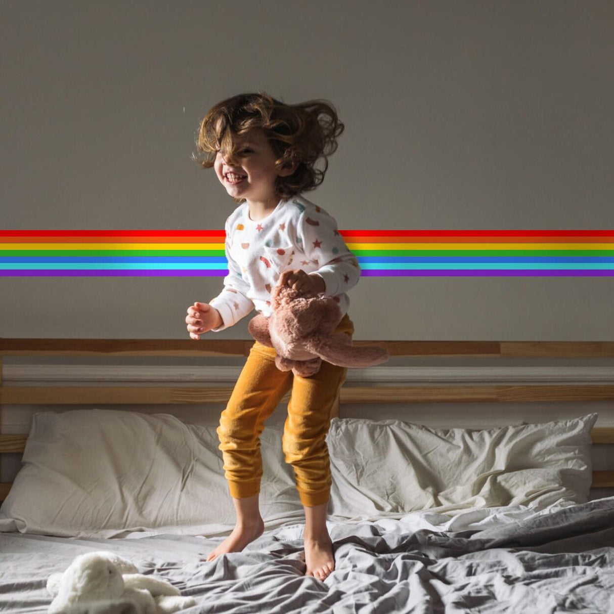 Happy child jumping on bed with rainbow stripe vinyl decal in nursery, holding a stuffed toy.