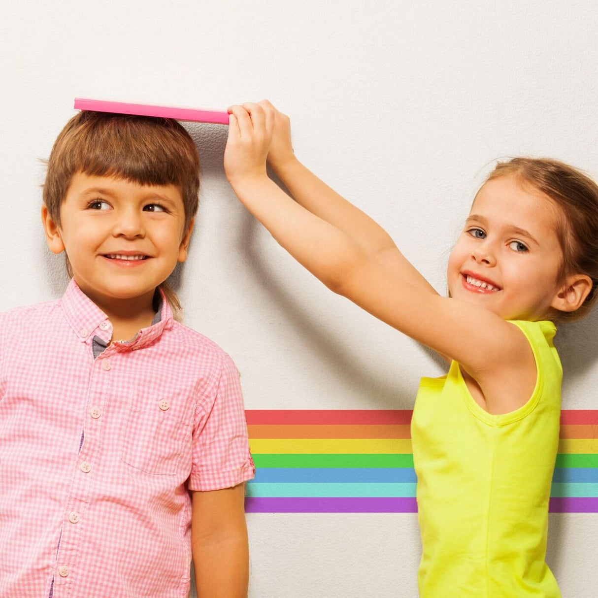 Two kids measuring height with a rainbow stripe sticker on the wall, perfect for nursery and birthday decor.