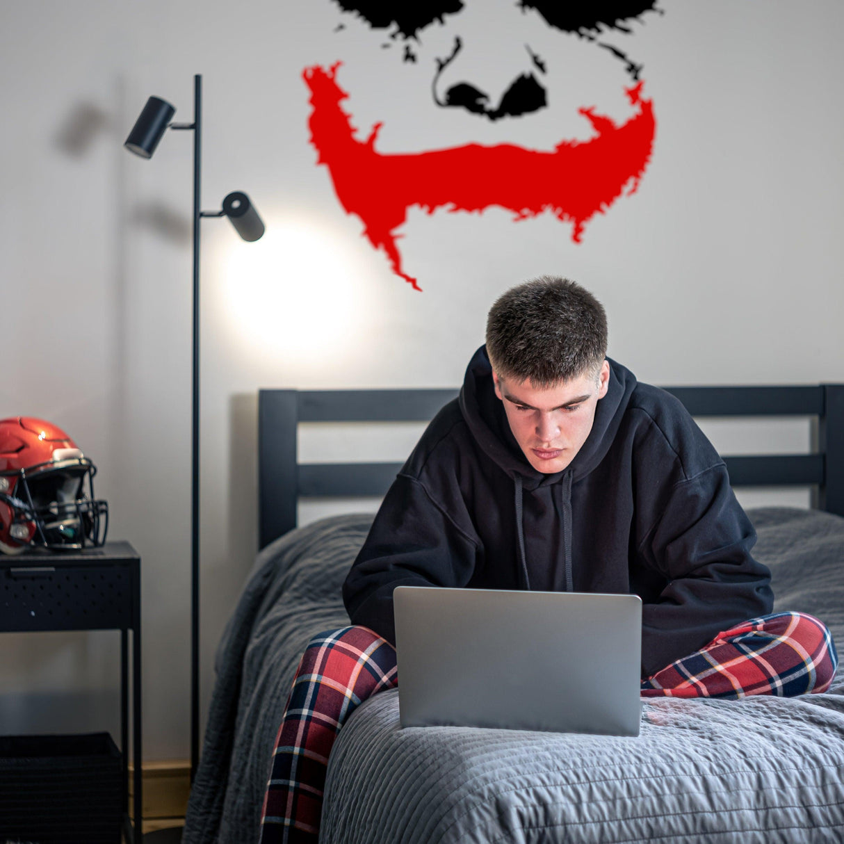 Young man sitting on a bed using a laptop under a Joker wall sticker with a red smile and black face.