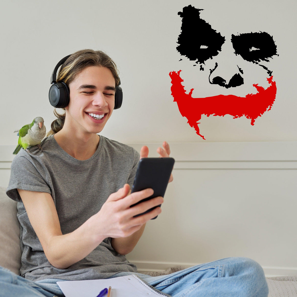 Cheerful young man with headphones smiles at his phone, featuring a Joker wall sticker in the background.