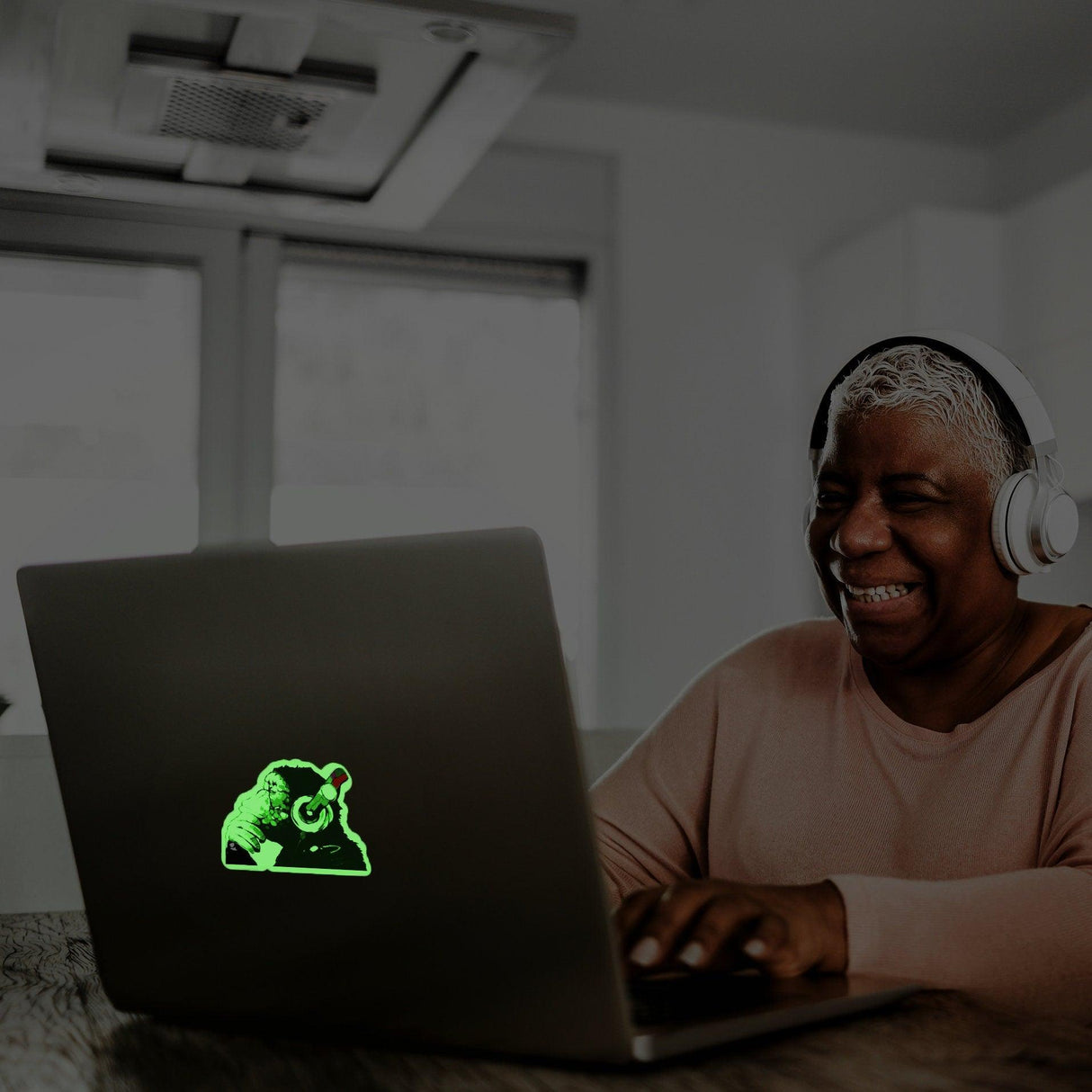Smiling woman with headphones using a laptop featuring a glowing monkey decal in dark room.