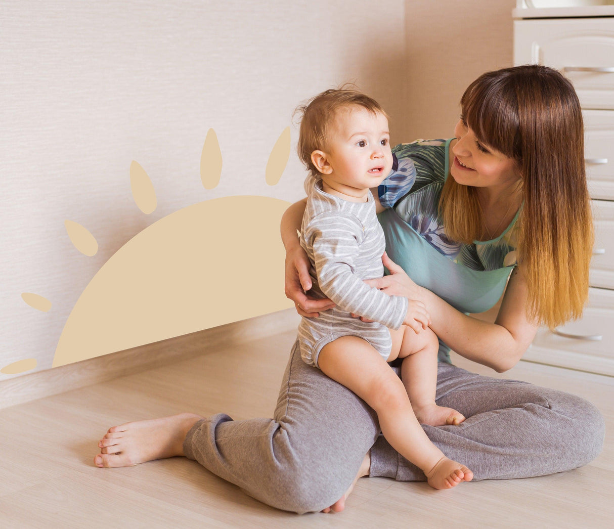 A mother sitting on the floor with her baby, with a large Boho sun wall decal in a nursery setting.