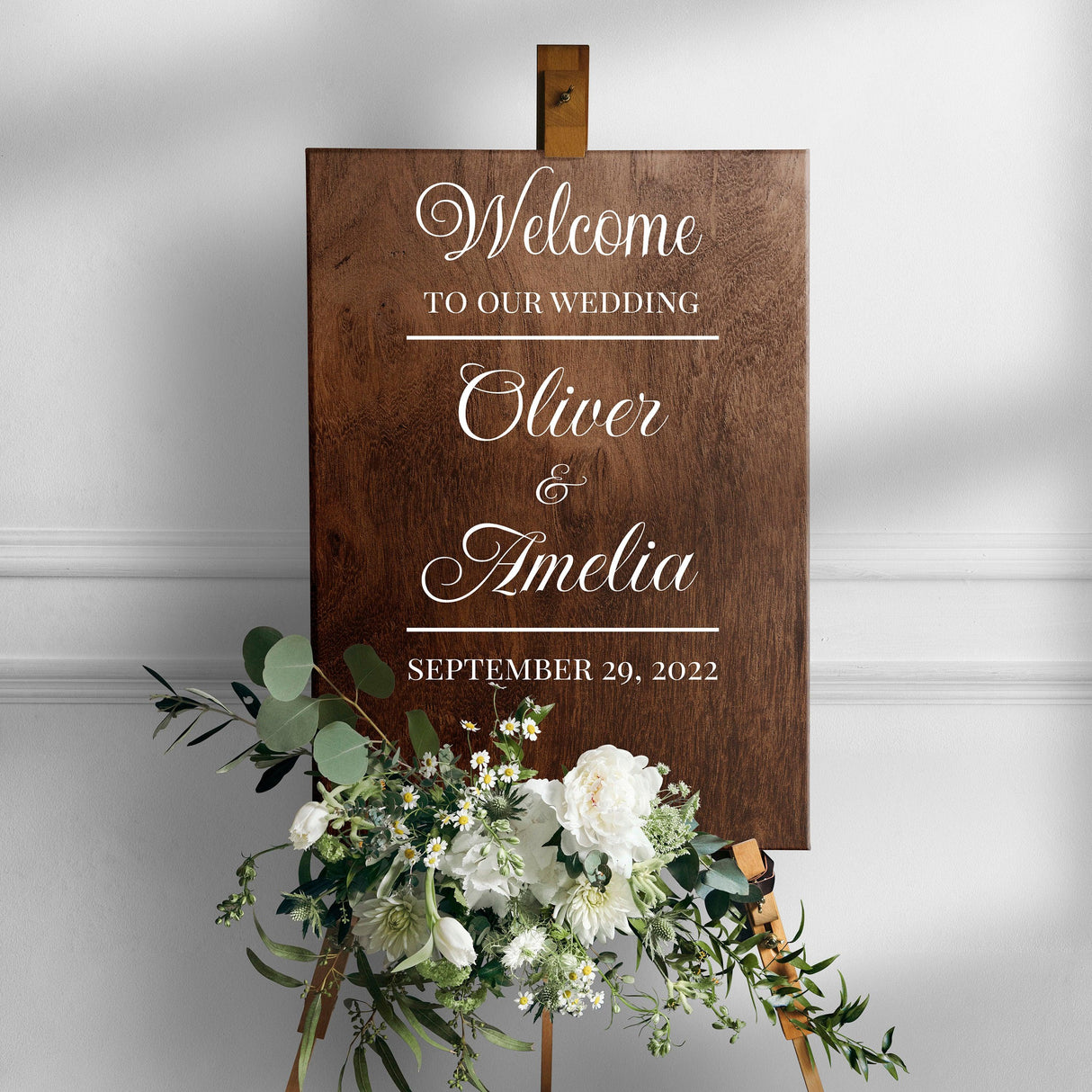 Welcome to our wedding sign featuring names Oliver and Amelia, adorned with flowers and displayed on an easel.