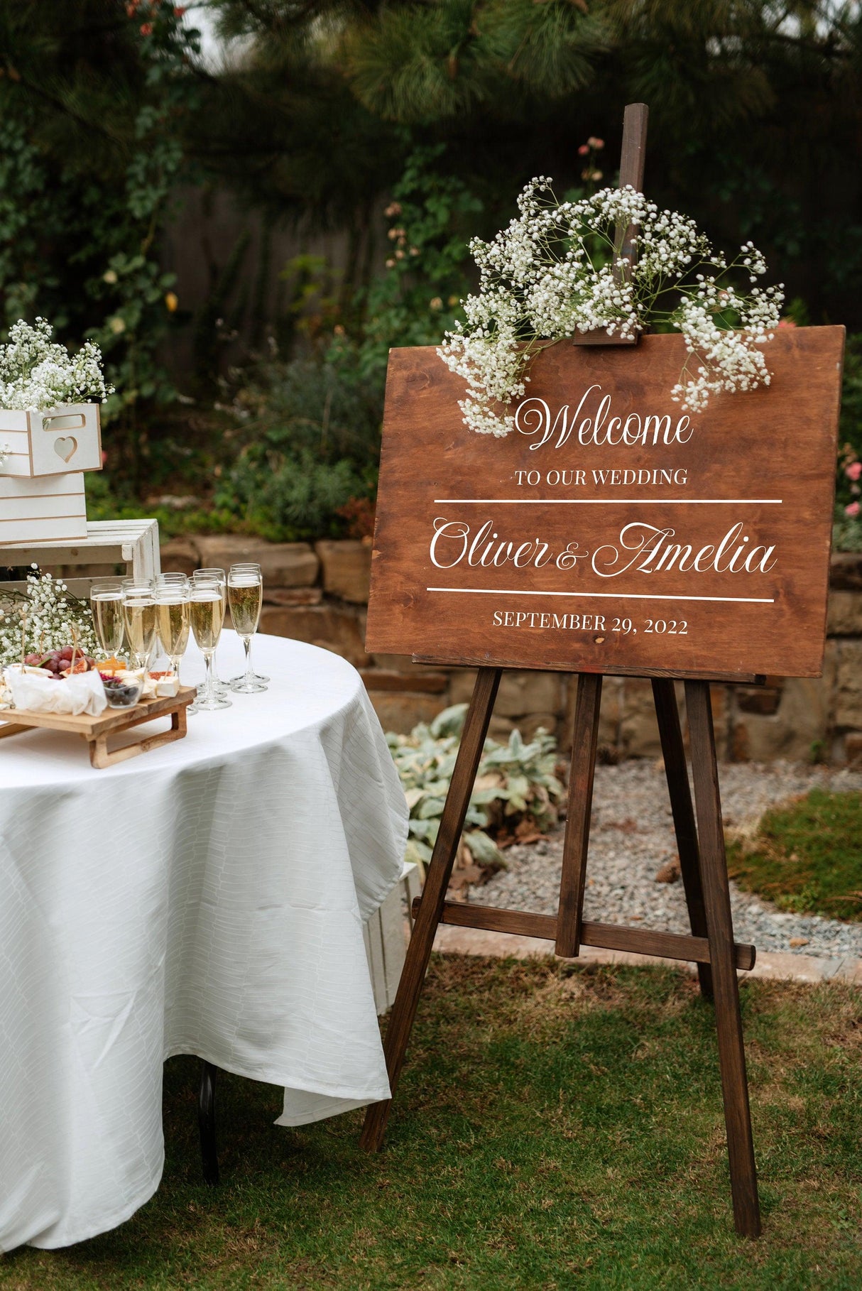 Personalized wedding welcome sign with names Oliver & Amelia and date, surrounded by flowers in a garden setting.