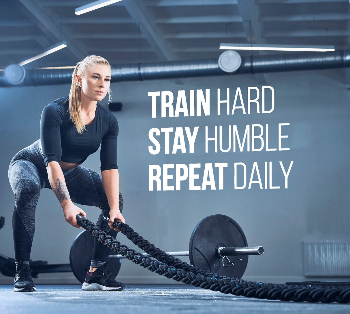 Motivational gym wall decal showing a woman working out with ropes, featuring the quote 'Train Hard, Stay Humble, Repeat Daily'.