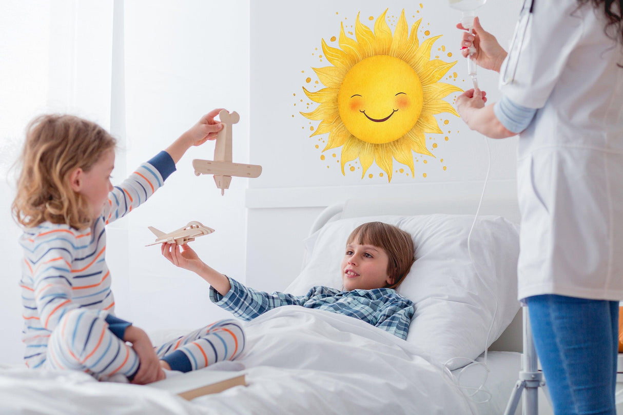 Two children play with toys in a hospital room, featuring a cheerful watercolor sun wall sticker for a warm atmosphere.