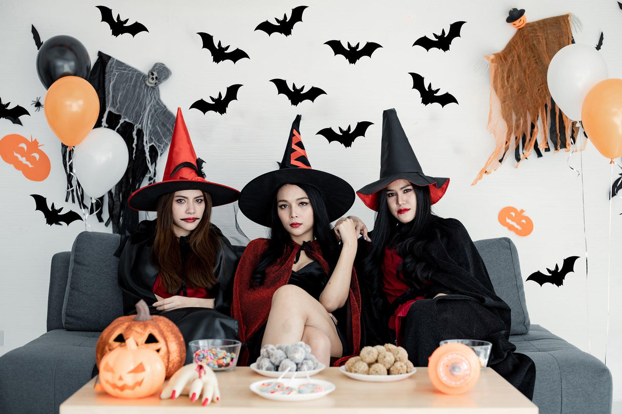 Three women in witch costumes celebrating Halloween with bats decorations and treats on a festive table.