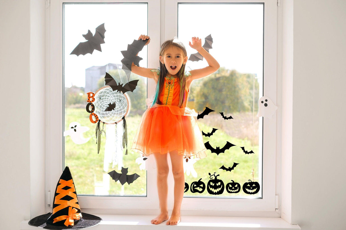 Happy girl in an orange dress posing by Halloween window decals with bats, pumpkins, and ghost decorations.