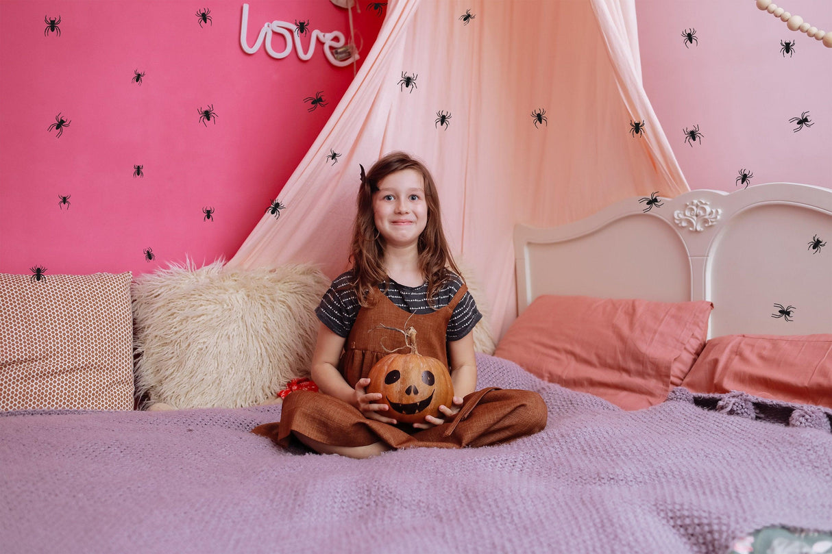 Girl sitting on a bed with a pumpkin, surrounded by creepy spider wall decals for Halloween decor.