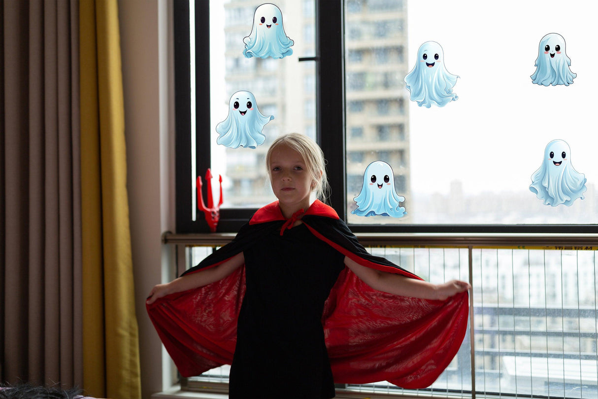 Child in a black costume with a red cape standing by a window decorated with white ghost stickers for Halloween.