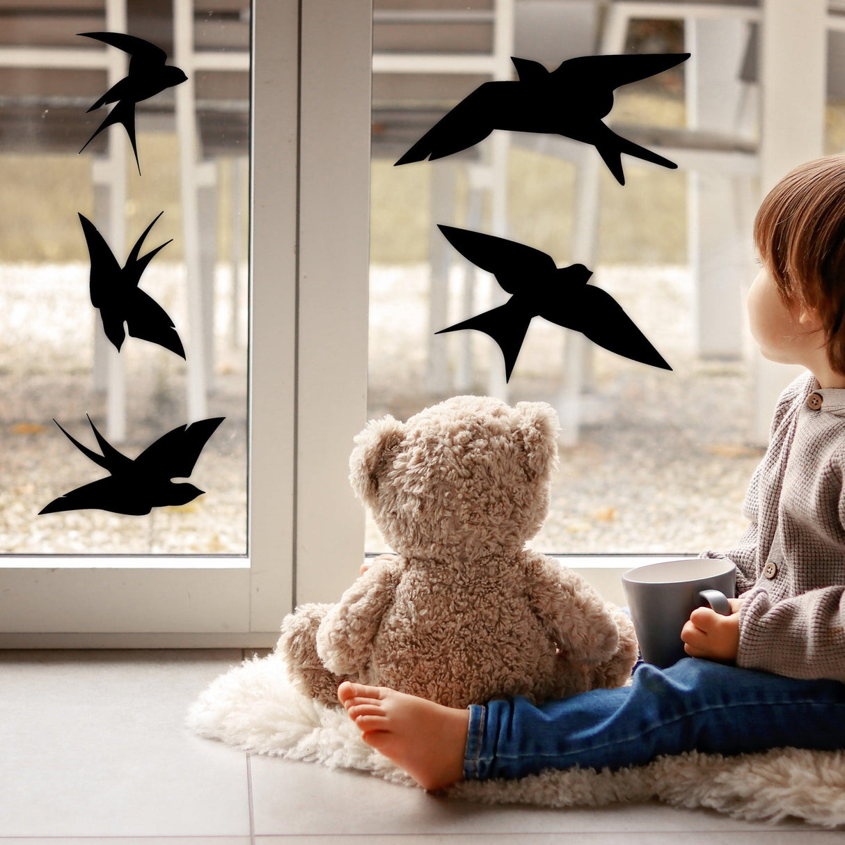 Child with teddy bear sitting beside windows featuring black bird deterrent decals to prevent bird collisions.