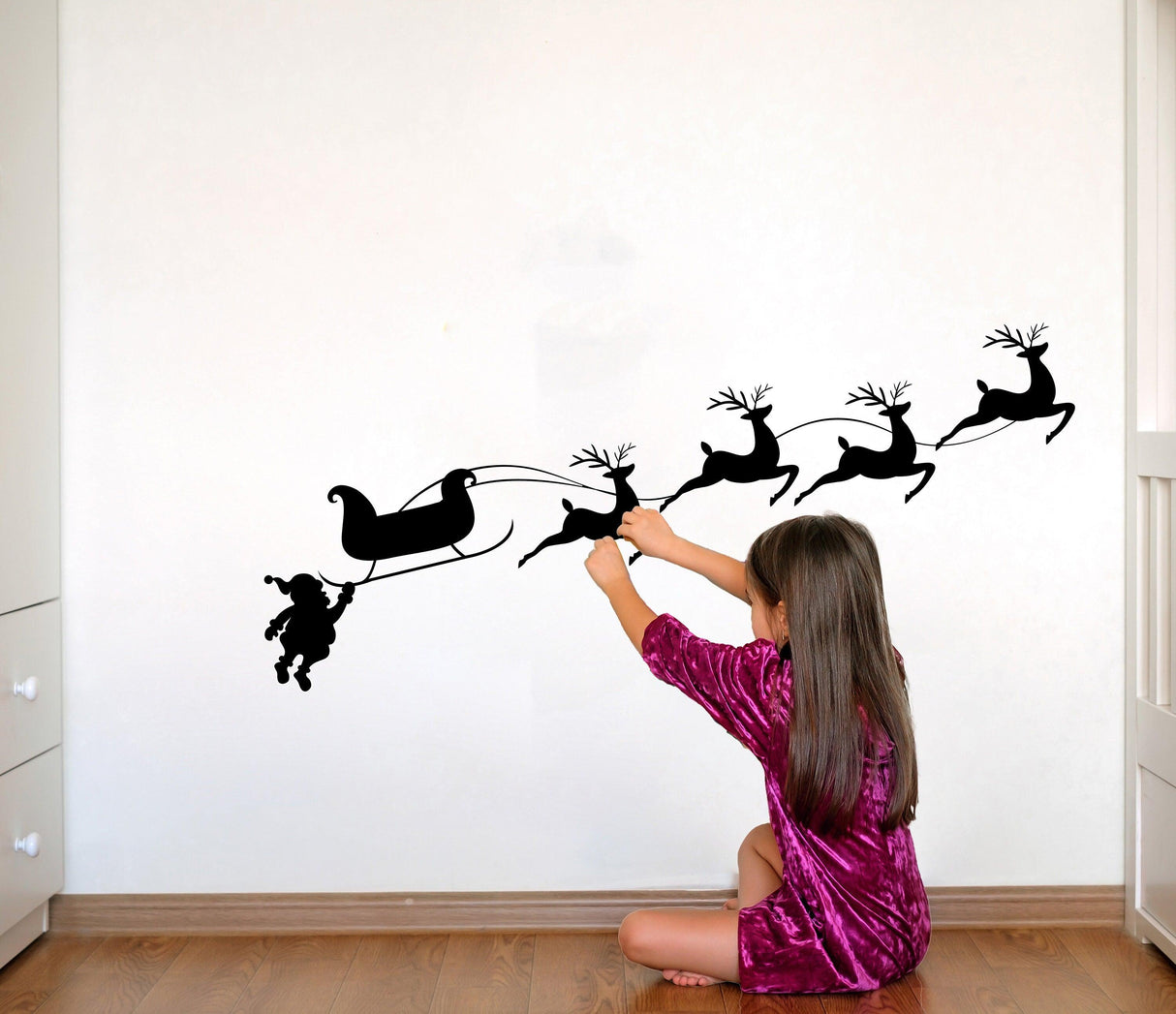 Girl in a pink dress playing with a humorous Santa and sleigh wall decal featuring reindeer silhouettes.