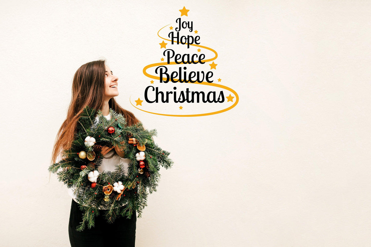 Woman holding a wreath next to a vinyl sticker with 'Joy Hope Peace Believe Christmas' on a wall.