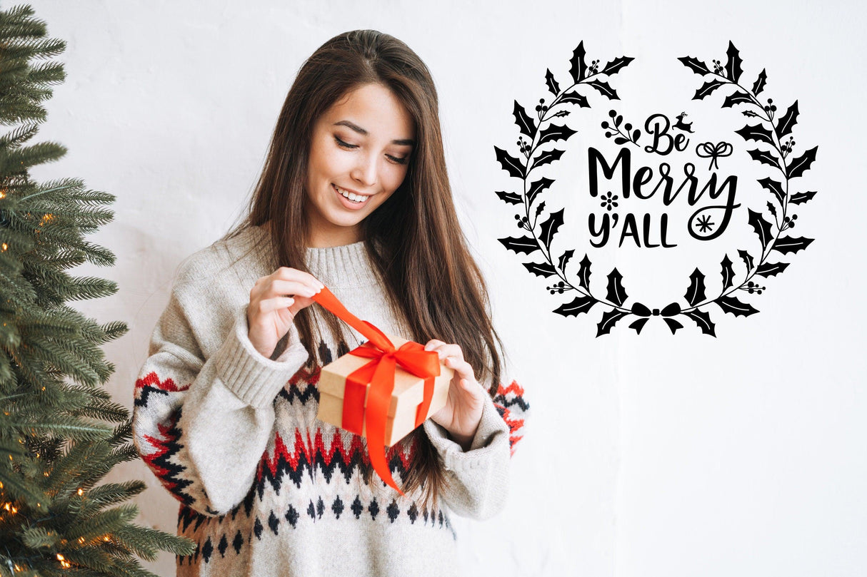 Woman in cozy sweater wrapping a gift beside 'Be Merry y'all' Christmas wall sticker.