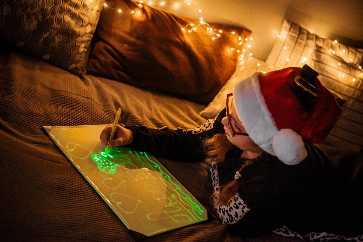 Child wearing a Santa hat draws glowing designs on a light-up drawing board in a cozy, festive setting.