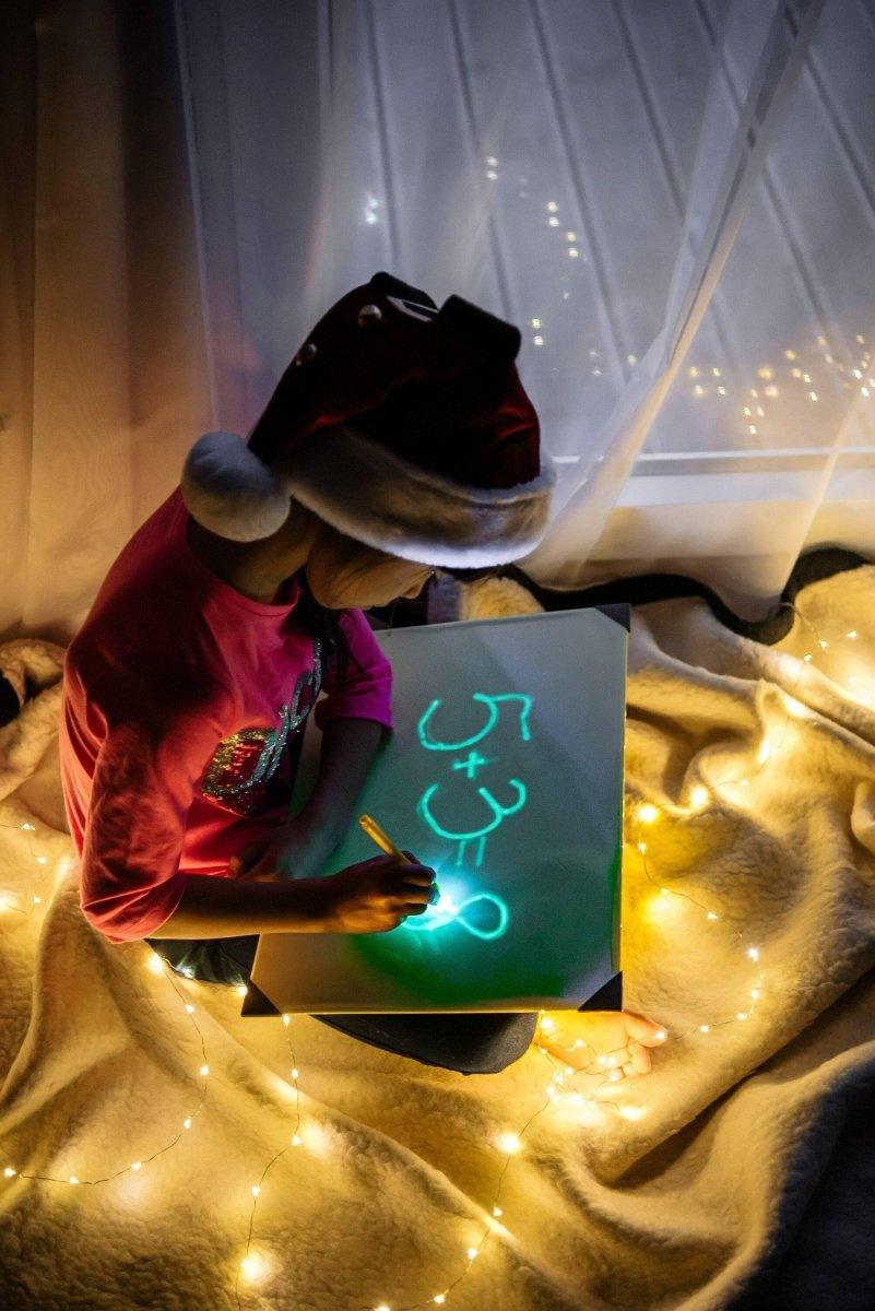 Child drawing glowing numbers on a light pad while wearing a Santa hat, surrounded by soft lights.