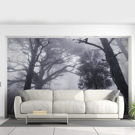 A dramatic misty forest wallpaper mural covers the entire living room wall, depicting tall, shadowy trees disappearing into fog. The grayscale tones create a moody, immersive atmosphere behind a modern beige sectional sofa and minimalist coffee table.