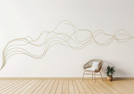 A large, abstract gold swirl line decal flows across a white wall in a minimal modern interior. A rattan chair with a white cushion sits below the decal, next to a small potted plant on a light wooden floor, creating a serene and contemporary atmosphere.