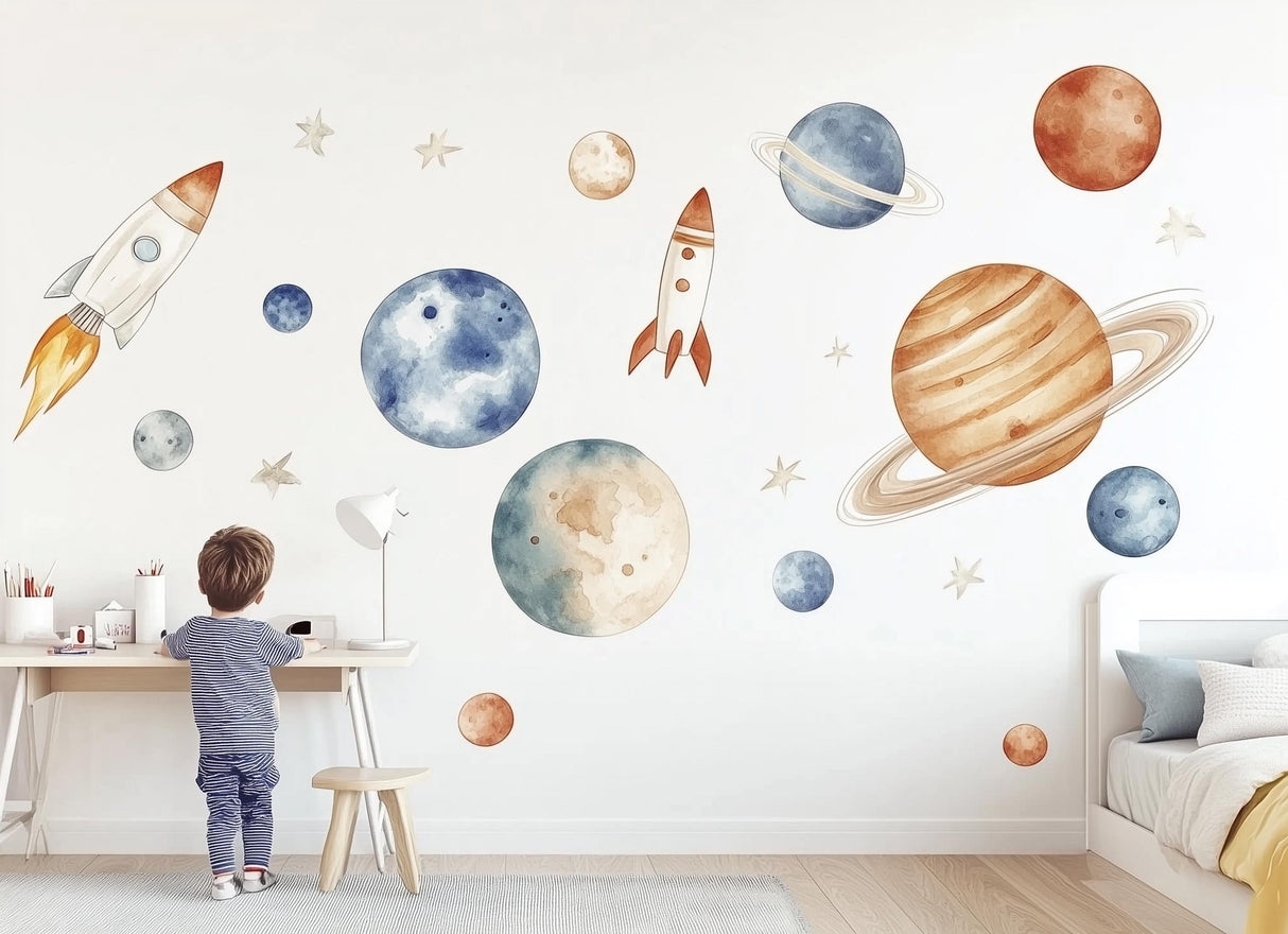 A child’s bedroom wall decorated with watercolor space-themed decals, including rockets, planets, and stars in blue, orange, and beige tones, creating a playful and dreamy outer space scene.