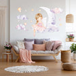 A whimsical wall decal showing a blonde fairy ballerina in a pink dress sitting on a crescent moon, surrounded by watercolor clouds and glowing stars, above a cozy white and pink-themed sofa in a nursery room.