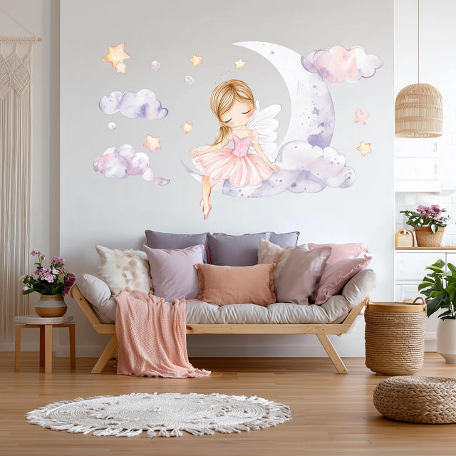 A whimsical wall decal showing a blonde fairy ballerina in a pink dress sitting on a crescent moon, surrounded by watercolor clouds and glowing stars, above a cozy white and pink-themed sofa in a nursery room.