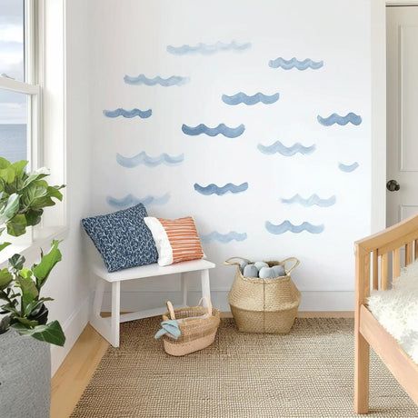 A set of blue watercolor wave decals applied randomly on a white wall in a coastal-themed kids&#39; room with a bench, pillows, natural baskets, and light wooden furniture.