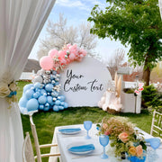 Outdoor wedding setup featuring a pastel balloon arch in pink, silver, and blue, arranged around a white backdrop arch.